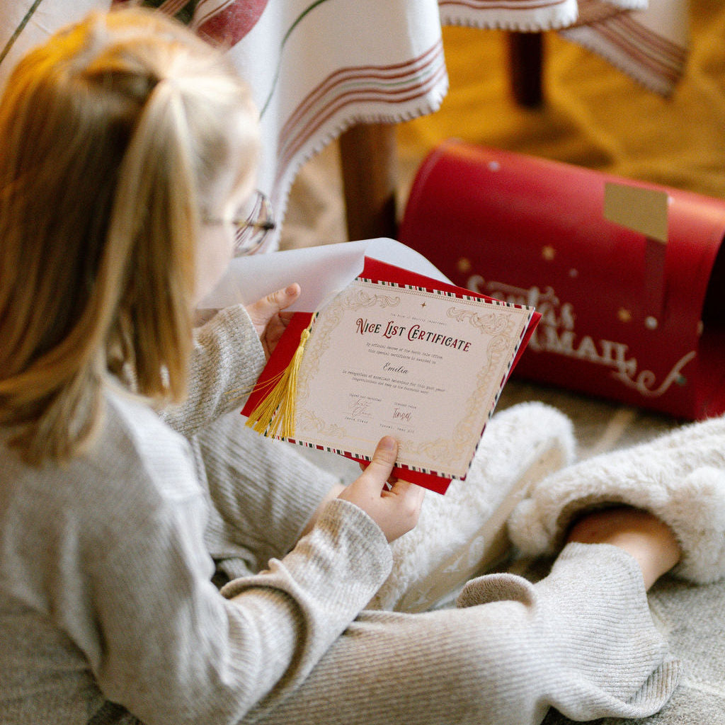 Child sitting on a carpeted floor holding a letter, with a striped blanket and wrapped gift in the background.